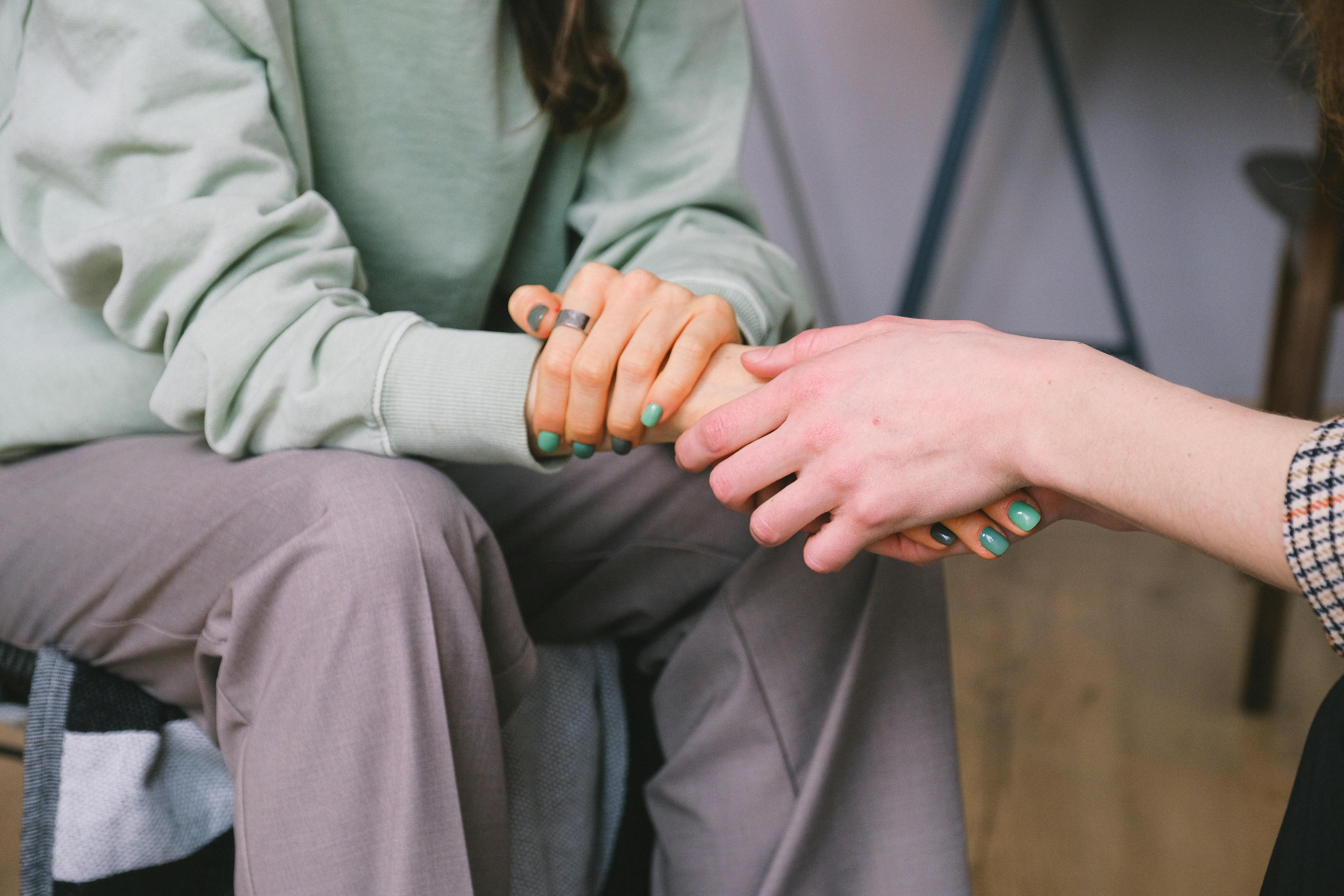 Two individuals are seated, one holding the other's hands in a comforting manner. Both have green nail polish, and one is wearing a plaid garment while the other is in a light-colored long-sleeve top and pants. The setting appears to be indoors with a casual atmosphere.