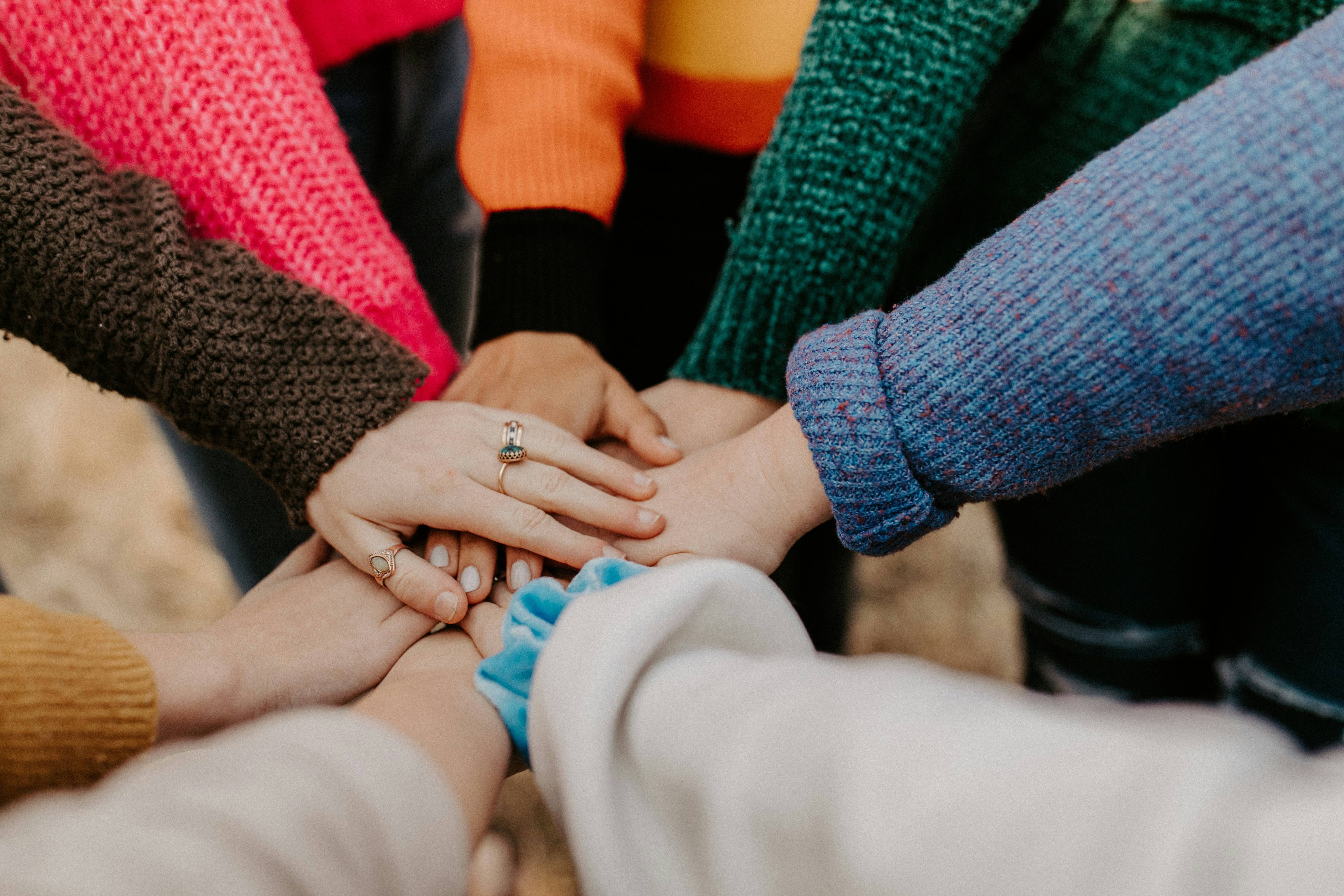 A group of individuals in colorful sweaters are standing in a circle, each reaching in to place their hand on top of the others in a gesture of unity.