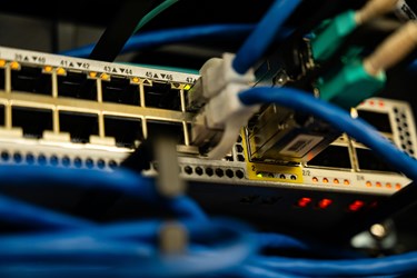 Close-up photograph of a network switch with multiple Ethernet cables plugged into numbered ports. Blue and black cables connect to the device, while green and amber indicator lights show active network connections in a data communications environment.