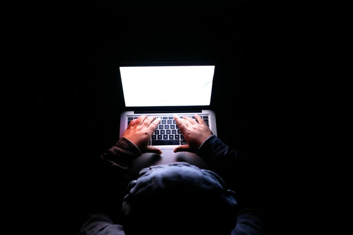 Person typing on a laptop in a dark room, illuminated only by the bright white glow of the screen, with hands positioned over the keyboard against a mostly black background.