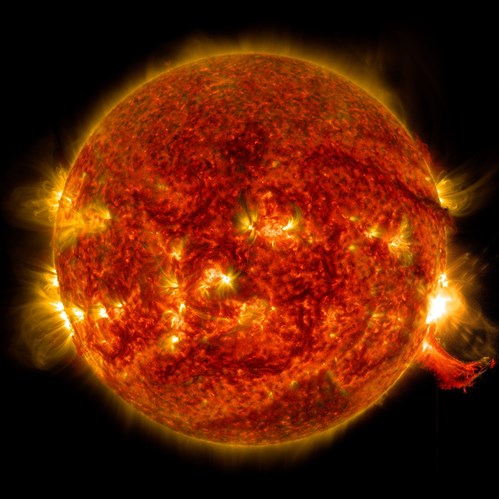 Full-disk view of the Sun glowing in deep red and orange tones, with multiple bright active regions and flares scattered across the surface, and a prominent burst of plasma extending outward along the right edge into the dark background of space.