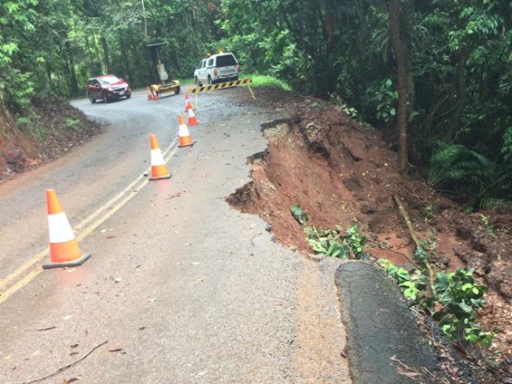 A rural road bordered by dense forest shows damage from a landslide, with part of the asphalt collapsed along the edge into a muddy slope. Traffic cones and a barrier mark the hazard, while vehicles are stopped nearby.