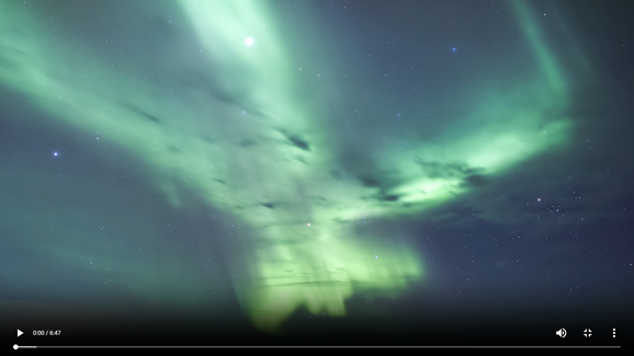 Green ribbons of the aurora borealis sweep across a dark night sky filled with stars. Bright, flowing bands of light stretch and swirl overhead, creating a vivid display of green and blue tones against the darkness.